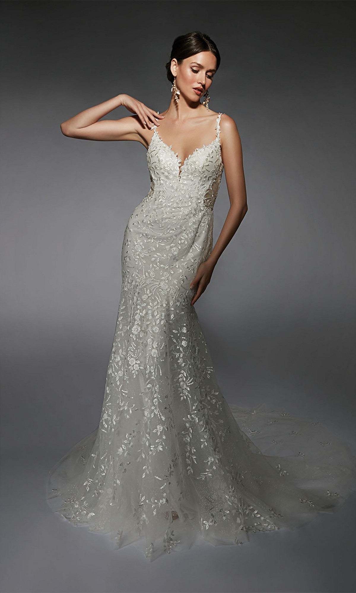 A woman poses in the Alyce 7122, a fitted long white dress featuring thin straps and a sheer train adorned with floral lace appliqués.